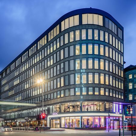 Yotel Глазгоу Екстериор снимка The photo shows a modern office building with a distinctive curved shape and a facade made of large glass windows. It is illuminated against a twilight sky, giving it a contemporary appearance. The ground floor features large glass entrance areas, li